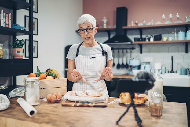  An older woman in an apron stands in a kitchen, explaining something as she's being filmed by a smartphone on a tripod. The woman is behind a large wooden counter with some flour-dusted dough on a tray directly in front of her. A crate of fruit, a container of sugar, and a trio of eggs sit to one side of the table, while a glass jug of milk and a plate of pastries sit off to the other side. The woman has short white hair and wears glasses, a white checkered apron, and a pale gray T-shirt.